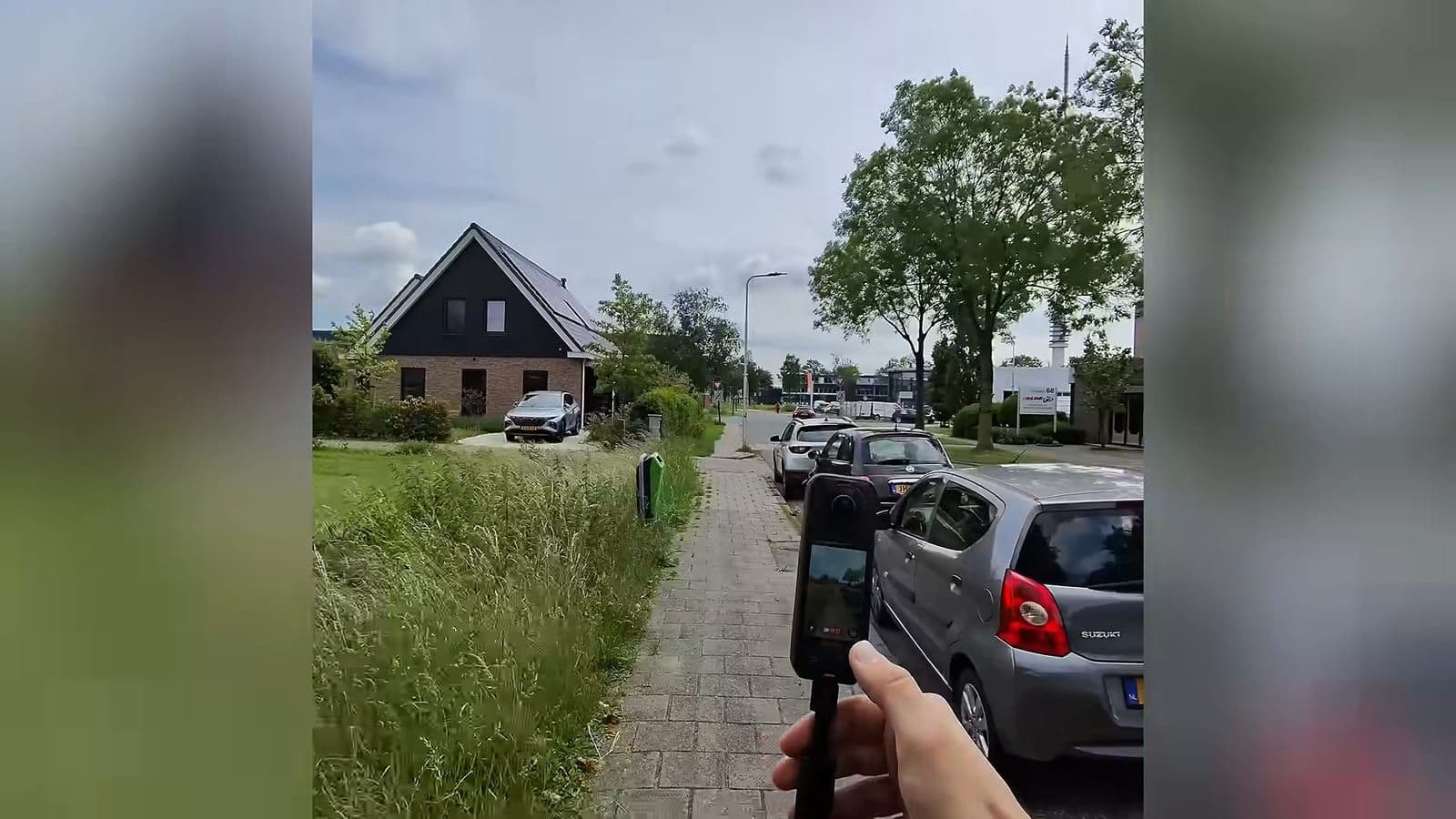 Hand houdt de Insta360 X5 op een korte selfie stick vast tijdens een wandeling over de stoep