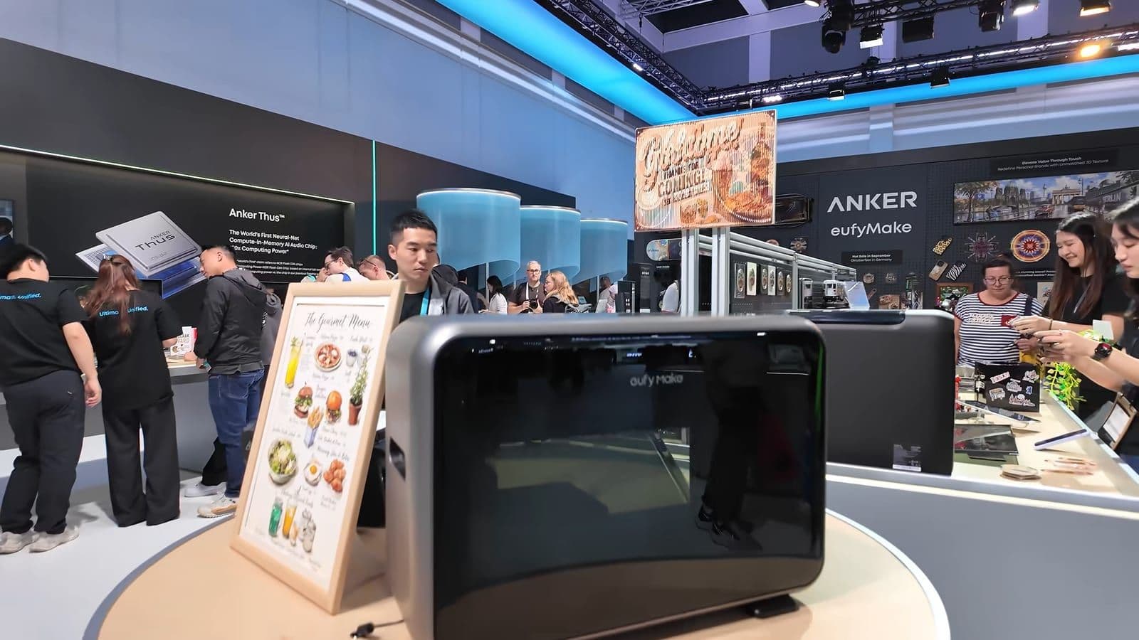 Zwarte eufyMake UV-printer op een ronde tafel op de Anker-stand, met bezoekers en voorbeeldprints op de achtergrond