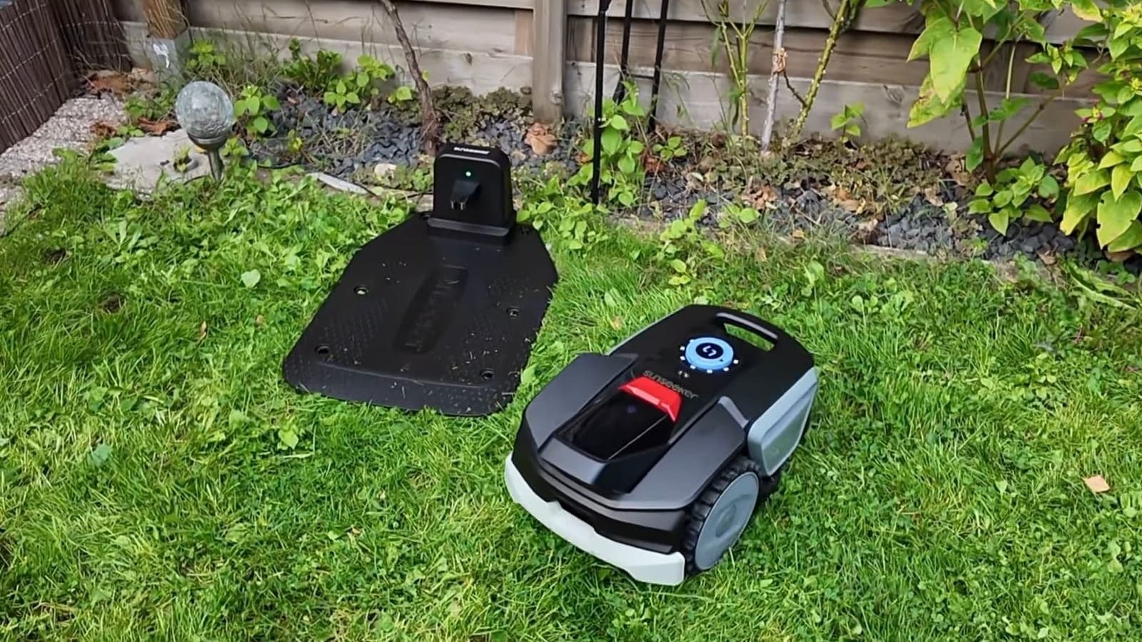 Sunseeker V1 robotmaaier op het gras naast zijn zwarte laadstation bij een border met planten