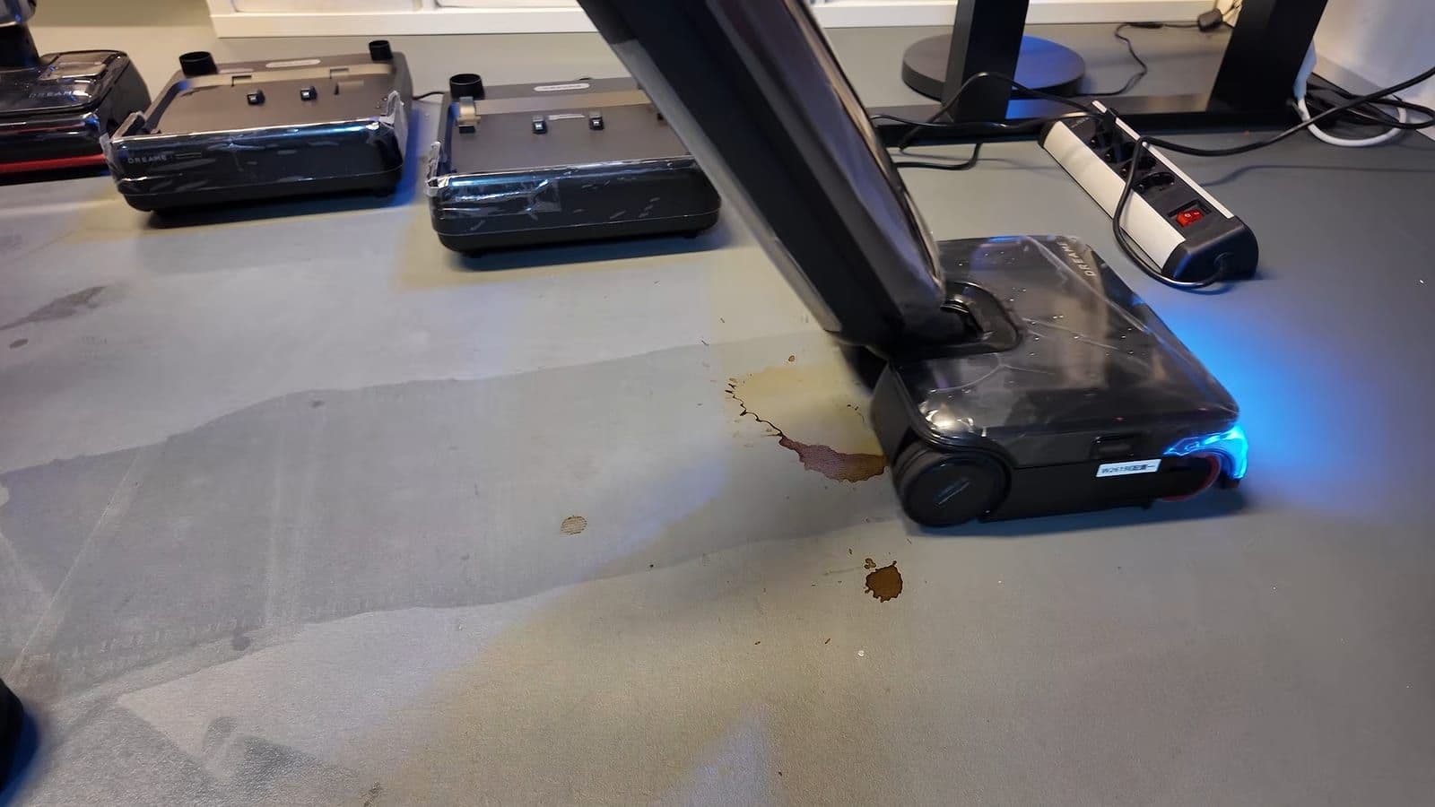 De Dreame T16 Pro Steam met blauwe verlichting in de kop rijdt over een opgedroogde bruine koffievlek op een grijze vloer